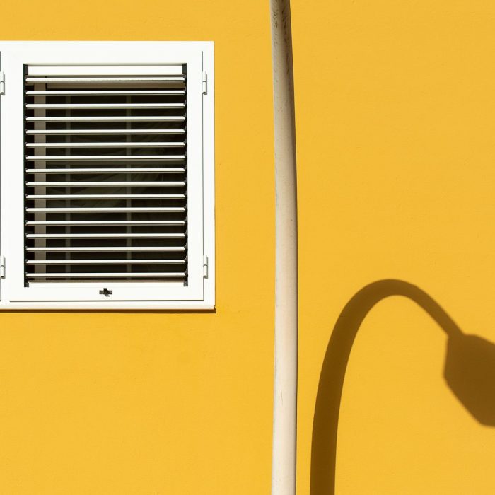 A minimalist scene featuring a yellow wall with a white window and the shadow of a lamp.