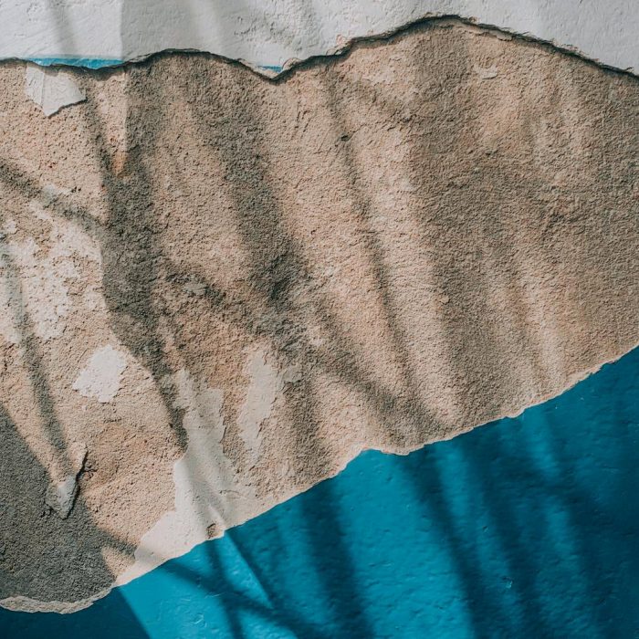 Detailed view of a wall with peeling blue paint and exposed plaster, showing textures and shadows.